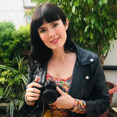 A woman with short dark hair and bangs wears a leather jacket and holds a camera