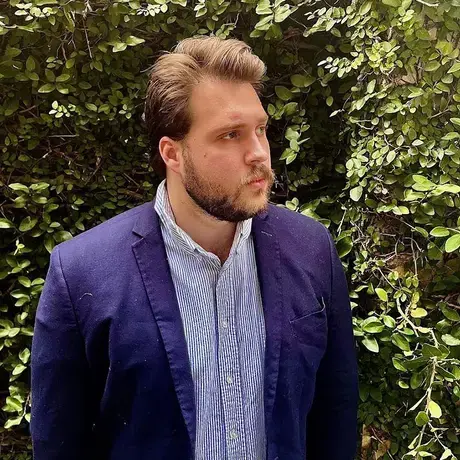 A man wearing a blue jacket and button down stands against a leafy background