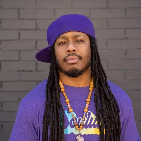 headshot of Montu Miller, who wears a purple t-shirt and hat