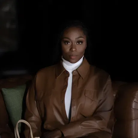 An image of Kayla seated and posed for a professional photo. She is a Black woman wearing a white and brown outfit..