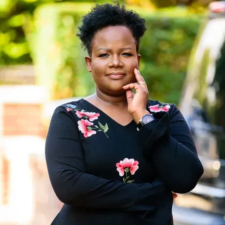 A professional photo of Norka. She is a Black woman wearing a black floral dress. She is posed with her hand touching her chin and has a blurred outdoor background.