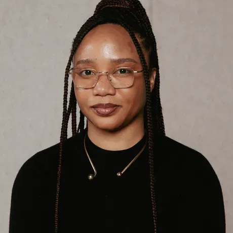 A headshot of Minne Atairu. She is wearing a black shirt and glasses. Her hair is in a long braided ponytail.