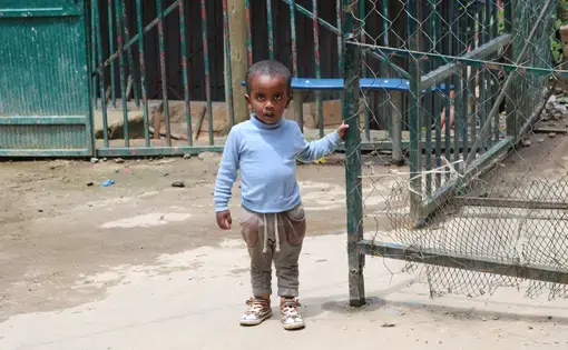 A young boy stands by the gate waiting to go inside to have lunch. He will spend his life in the orphanage unless he is locally adopted. Image by Abigail Bekele. Ethiopia, 2018.