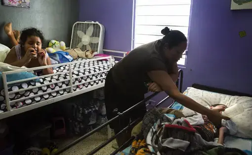 Karen Feliciano, center, feeds her oldest child, Kenny, 18, who suffers from a number of neurological and physical conditions, while her youngest daughter Kylia, who suffers from autism plays on the bed in their home. “When [Kenny] was born, the doctors said he would only live to six,” Karen said. Image by Ryan Michalesko. Puerto Rico, 2017.