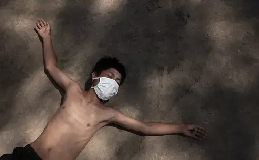 A young boy lies down on the dirt ground with arms outstretched. He is wearing a white surgical mask, black shorts, and no shirt.