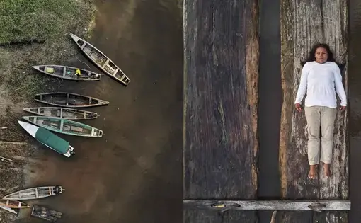 To the left, a series of empty wooden canoes stand by the margins of a river. On the right, Lilia Isolina Java Tapayuri lies down on a plank of wood, looking directly at the camera, captured from an aerial shot. She is wearing a white long sleeved shirt, khaki-colored pants, and no shoes.
