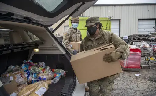 People in medical masks load boxes into the trunk of a car.