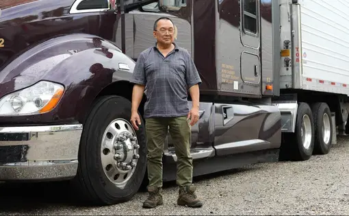 An older man wearing glasses, a blue striped shirt, olive pants, and boots stands in front of a black semi truck.
