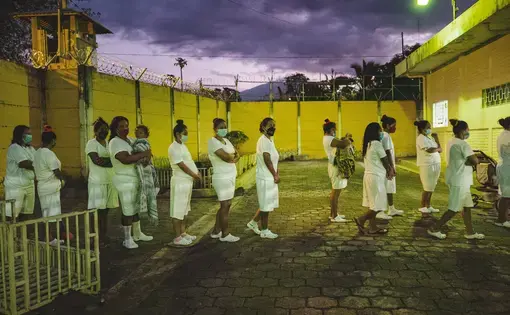 Waiting to go inside, a line of women stand in a yard enclosed by a wall that is topped with barbed wire. The women wear the same all-white uniform of a t-shirt, shorts, and crocs, and they all wear masks. Two women are holding small children. On the right of the frame is a stroller.