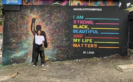 A woman stands in front of a mural with her fist raised. Her pose is echoed by a figure in the mural. Text on the wall reads, "I am strong, Black, beautiful, and my life matters."