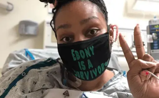 A selfie of a woman in a hospital bed making a peace sign. Her dreadlocks are in a bun at the top of her head and she wears a black face mask that says "Ebony is a survivor" in green.