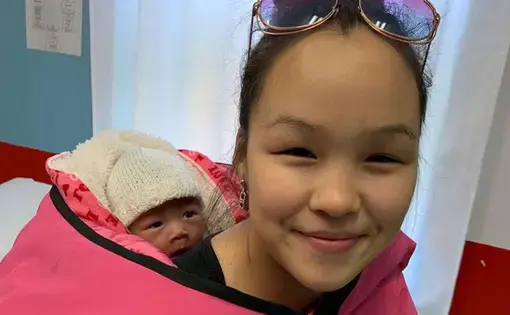 Image of a smiling young woman with an infant secured on her back with a pink blanket. The woman has sunglasses propped on her head, and the infant is wearing a white knit hat.