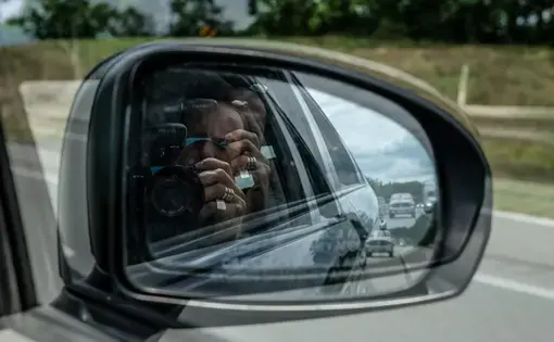 Self-portrait of a man taking a photo of himself with a DSLR camera in the passenger side mirror of a car. A double exposure effect makes the image look slightly blurred.