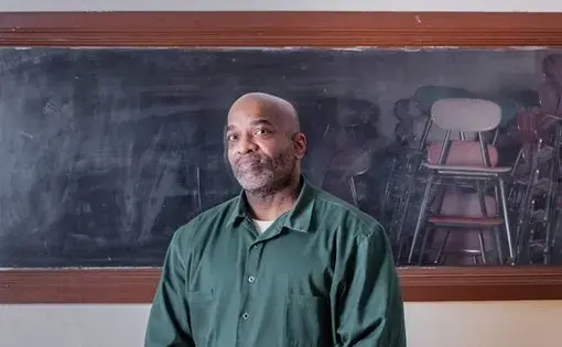 Portrait of a man wearing a green prison uniform standing in front of a horizontal window. Stacks of chairs are visible on the other side of the window.