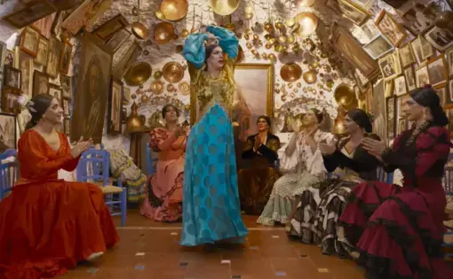 A man in drag wearing a blue and gold dress dances in the middle of a room. Sitting around the perimeter of the room and clapping along are five other men in drag. The walls are covered in gold decorations and framed pictures.