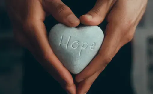 A pair of hands holding a heart-shaped stone that reads "Hope"
