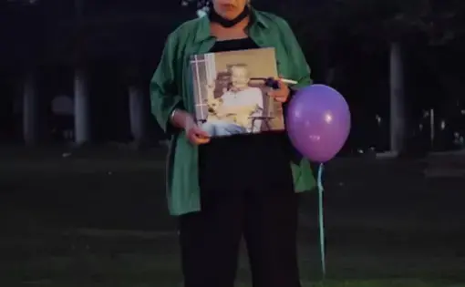 a woman holds a portrait of her deceased friend, three candles are on the ground in front of where she stands in a park at dusk. she has a purple balloon.
