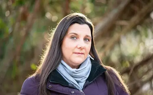 A woman wears a scarf and purple coat in front of trees