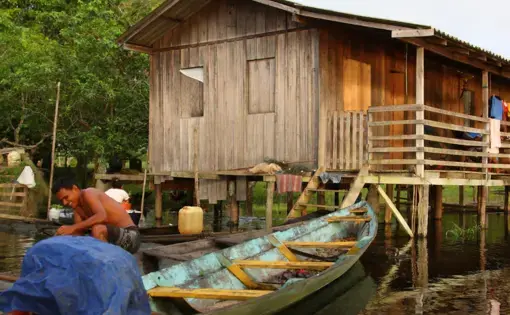 A person paddles a boat. In the foreground, there is a wooden shack on stilts.