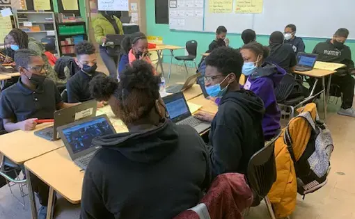 Black students, ages 12-13, in dark blue uniforms and wearing masks turn towards each other to have a discussion