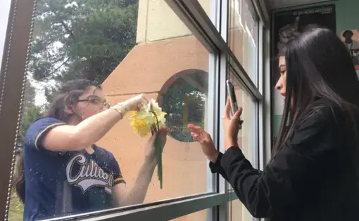 One high school student stands outside a classroom window holding up a yellow flower, while another student stands inside the classroom taking a picture through the window on their phone.