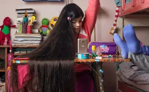 Photo of a young girl sitting at a desk in her bedroom raising her arm as if to ask a question. She has long black hair draped over the desk.