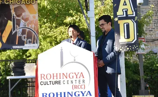 a child of Rohingya origin speaks at a podium at an event alongside a young adult of Rohingya origin. They both wear suits.