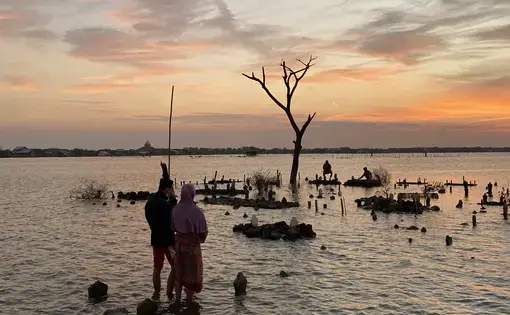 People stand in a flooded cemetery on the edge of a large body of water as the sun sets.