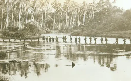 Black and white photo of WWII-era soldiers wading across a shallow body of water single file.
