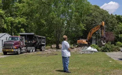 A woman stands in a field observing the destruction of a house in the background by a bulldozer.