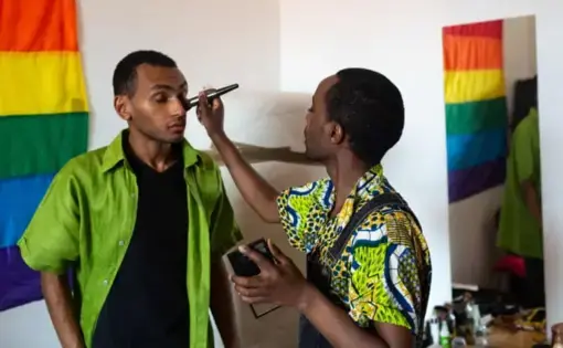 One person uses a brush to put makeup on another person's face. On the wall behind them, a rainbow flag hangs.