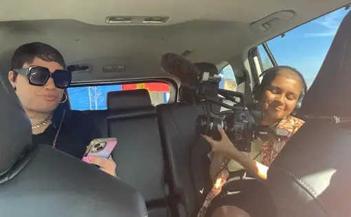 Two individuals sit in the back of a car, one of them is holding camera equipment.