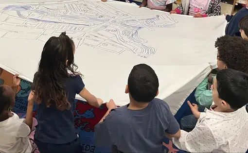 Kindergarten and first grade students sit around a large paper with a drawn map of Lowell, MA