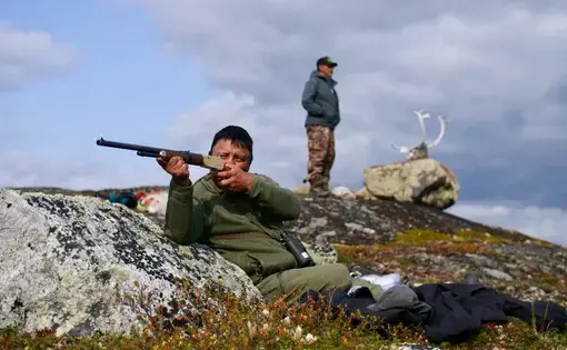 A man sits against a rock aiming a hunting riffle