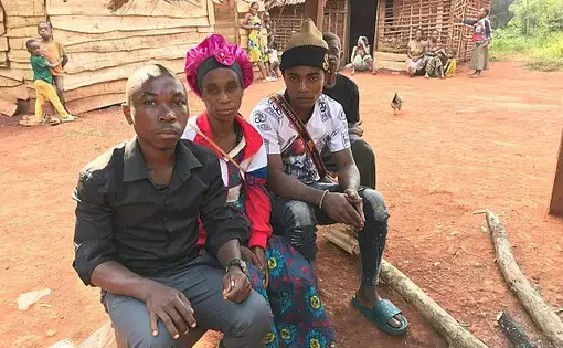 Three Baka individuals sit on a bench outside a wood and clay building