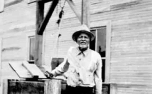 Portrait of Nap McQueen, an elderly Black gentlemen taken around 1936. It is a black and white image in which Nap in black trousers, a collard shirt, and hat stands in front of a well