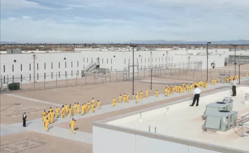 An aerial shot of of a prison yard. A line of inmates in yellow walking while being observed by prison guards.