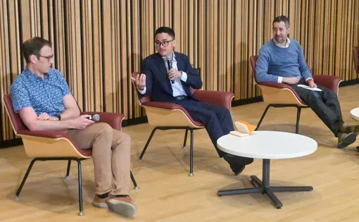 Three men sit in chairs, one with microphone talks