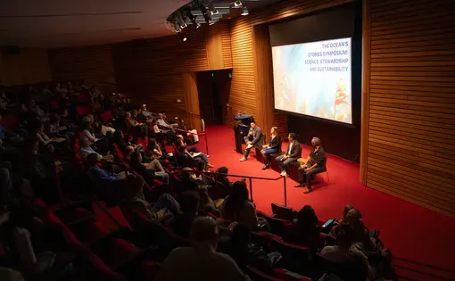 Four man sit for a panel in front of screen in auditorium