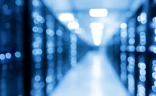 A blurred image of a dimly lit datacenter hallway showing out of focus vertical stacks of lights emitted by computer processors.
