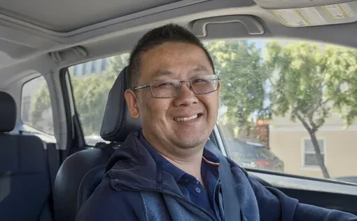 A smiling man wearing a blue sweater and blue shirt smiles while sitting in a car.