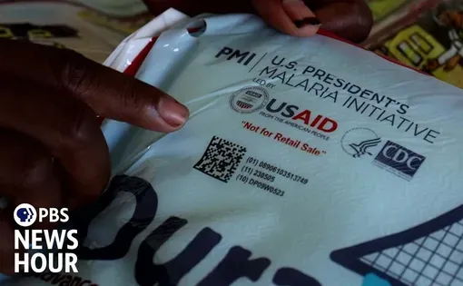 a person's finger points to the labels on a USAID aid package for malaria. PBS NewsHour logo. Video thumbnail for "Ghana struggles to fight disease and poverty without vital U.S. aid"
