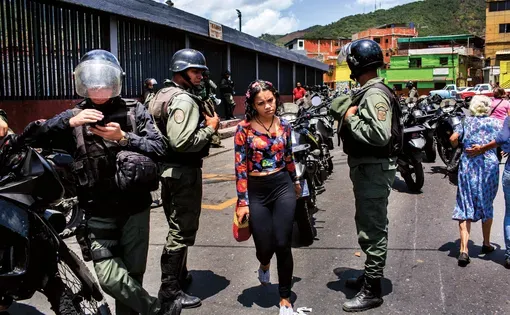 A young woman looks directly at the camera as police in military gear surround her, with colorful buildings in the background