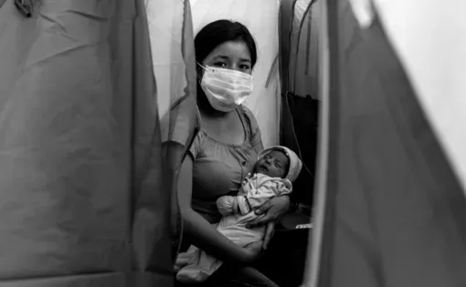 a woman wearing a face mask holds a child in her arms in a makeshift outside structure