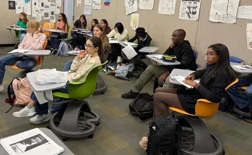 Students in a highschool classroom sit listening to a news report with writing packets on their desks
