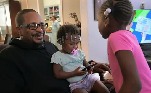 Cecil Burton rocks his granddaughters Chasity Shufford, 1, and Katelyn Ellis, 6, during a family gathering while their mother, Keisha, cooks fried chicken in the kitchen. Burton, 54, said child support once garnished so much of his paycheck he didn't have enough to live on. His coworkers took up a collection for him. Image by Karl Merton Ferron / Baltimore Sun. United States, 2019.
