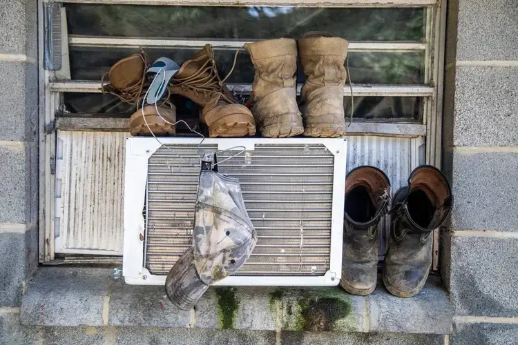 Boots and a hat dry on a window air conditioning unit at a Johnston County farmworker camp Thursday August 27, 2020. Air conditioning is considered a luxury by many farmworkers in North Carolina farmworker camps.&nbsp;Image by Travis Long. United States, 2020.