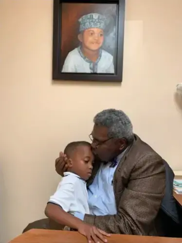 Marcos A. Rivera hugs his young grandson, who is also pictured in a painting above his office desk. Image by Natasha S. Alford. United States, 2019.