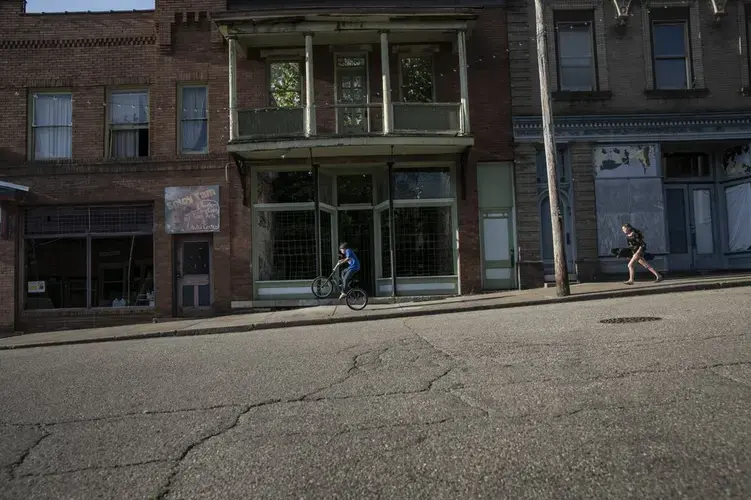 A boy rides his bicycle while a girl runs with her skateboard past abandoned shophouses along the streets of Shawnee, Ohio. Image by Wong Maye-E/AP Photo. United States, 2020.