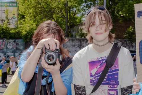 two teenagers face the camera, the one on the left holds a camera in front of their face.